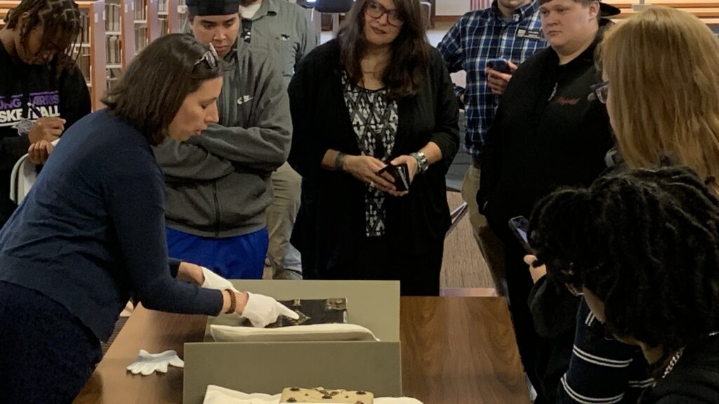 People look down at a book with metal fittings on its cover, while one woman wearing gloves points to those fittings