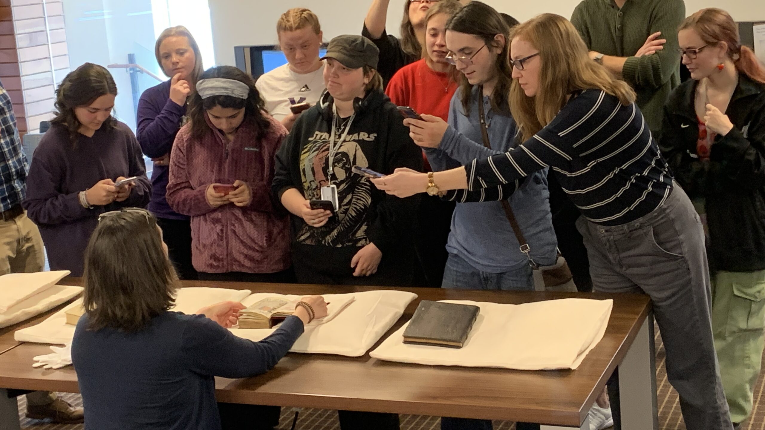 Students lean over a desk with their phones open to shoot photographs. On the desk is an open medieval manuscript