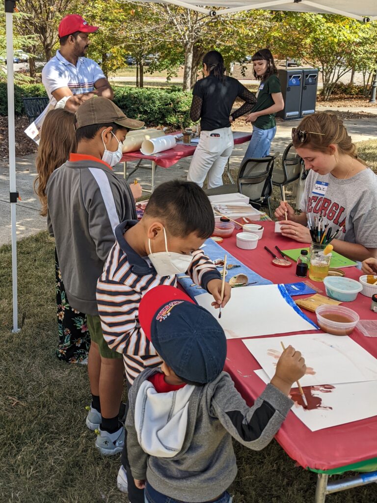 A row of children stand in front of a table outside, holding paintbrushes and painting. Student helpers stand in the background.