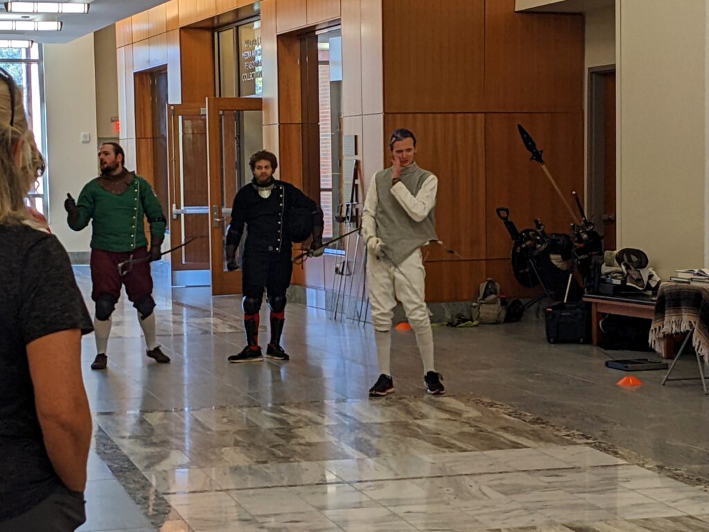 Three men in padded jerkins stand in a modern indoor space, holding swords.