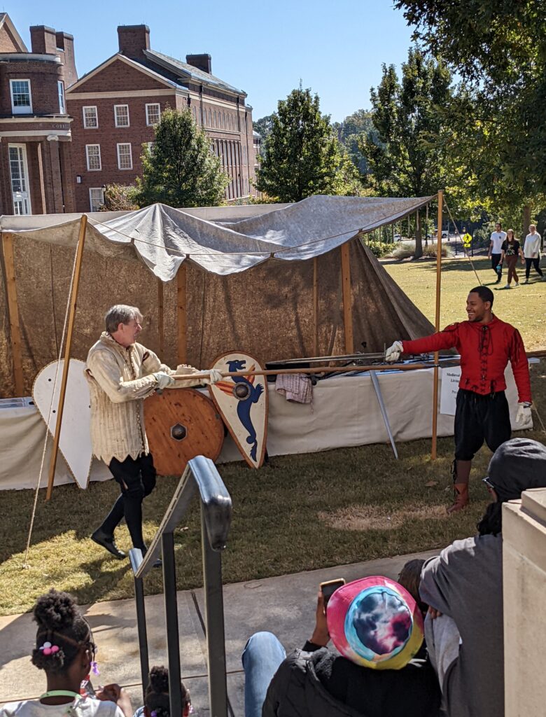 Two men in medieval clothing stand under a canopy in front of a table holding medieval weapons and shields. They point medieval weapons at each other.