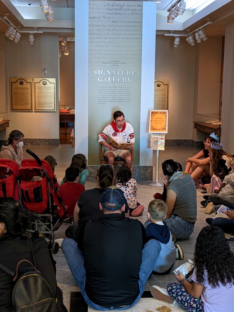 One student reads from a book while parents and children sit on the floor and listen.