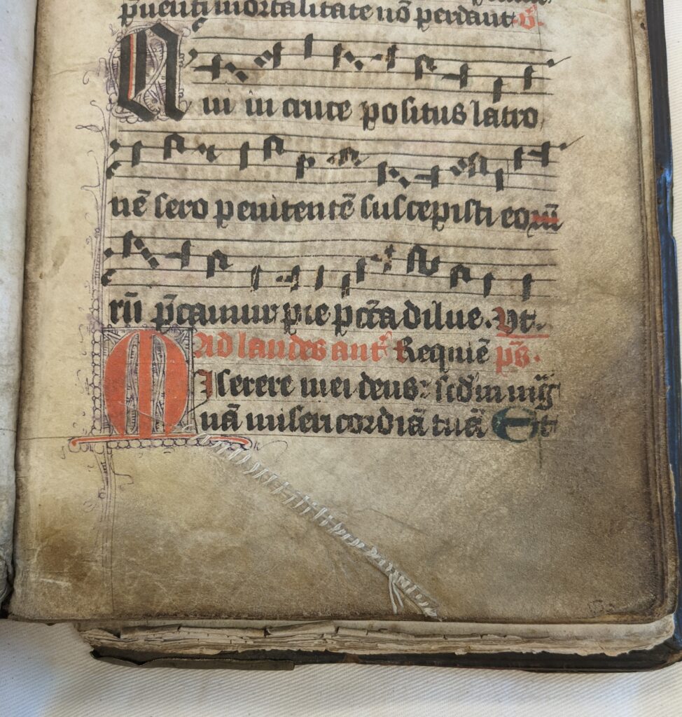 The bottom half of a worn parchment page written in red and black gothic script. Musical notation on 4-line staves is also present. There is a tear in the bottom margin of the page that has been sewn up with white thread.