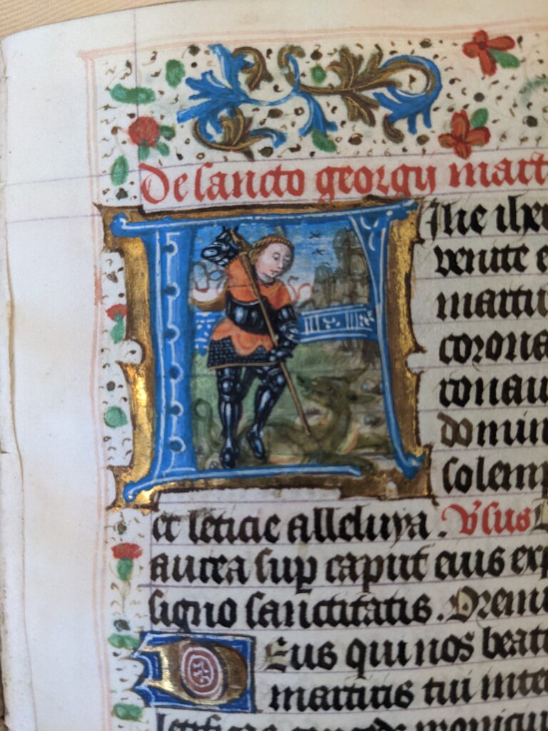 Part of a page, highly decorated, from a medieval book. On the page is a picture of a knight stabbing a dragon