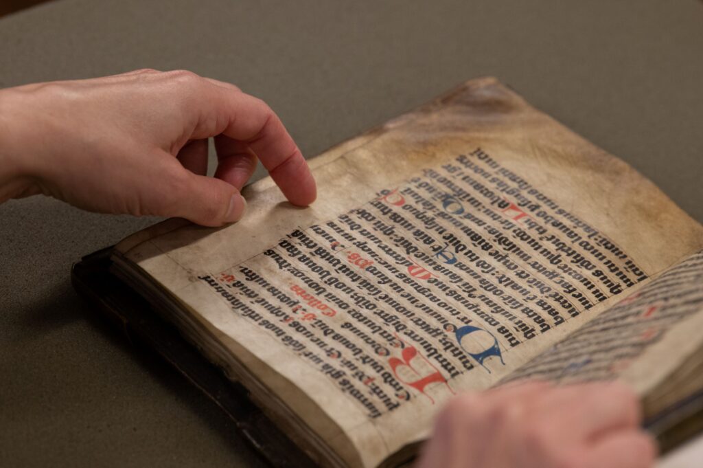 A book with dark lettering and colorful initials, the edges of its pages are very dirty.